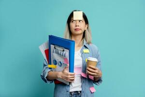 Bored and confused young Asian woman holding document file and notes on her forehead. She stands holding mug of hot coffee and looking sideways isolated on blue background. photo