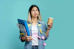 Asian female office worker is bored with a lot of paperwork at hand. She looks up, standing isolated on a pastel blue background. photo