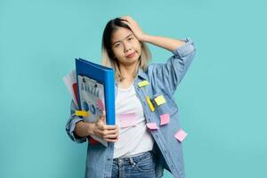 Asian female office worker holding head in hands and holding many document file standing isolated on blue background in studio. She was confused and had a headache. photo