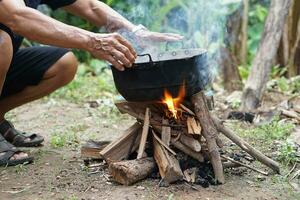 Close up man holds old black pot to cook on bonfire. Concept, cooking outdoor, kitchen in forest. Survival life skill for camping or hiking. Rural traditional lifestyle. Lighting fire with wood. photo