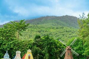 verde montañas adornado con lozano verde árboles, debajo un cielo pintado con sombras de sereno azul y vibrante turquesa nubes el fondo blanco imagen muestra viento turbina cuchillas multicolor edificio foto