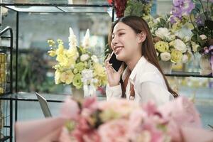 uno joven asiático hembra florista dueño, trabajando con computadora portátil, de venta floral arreglar, hablando en móvil teléfono en vistoso flor tienda Tienda con un hermosa manojo de flores, y comercio electrónico negocio. foto