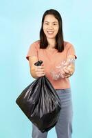 smiling asian housewife one hand holds plastic bag,other hand holds used plastic bottle on blue background,concept of waste management,campaign to separate household waste, support garbage recycling photo