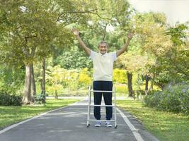 senior man with walker raising hands gesturing happiness while practice walking outdoors in the park photo