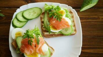 fried toast with lettuce, egg, cucumbers and red fish in a plate, on a wooden table. video