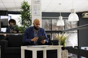 Arab businessman using tablet for digital work in business office. Company man employee scrolling through social media during break while sitting on couch in coworking space photo