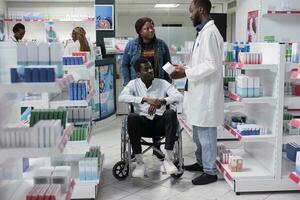 African american man in wheelchair and social worker choosing prescription treatment in drugstore, talking with pharmacist. Chemist recommending medicaments to client with disability, all black team photo