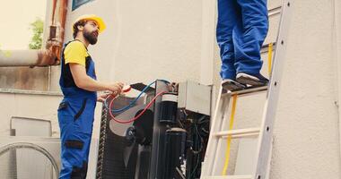 Proficient expert holding electric power drill stepping down from folding ladder after finishing servicing rooftop hvac system while coworker assembles manifold gauges to check freon levels photo