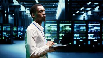 Administrator walking through server rackmounts delivering web content to clients over the internet. Manager in high tech facility using laptop to make sure critical systems are in faultless condition photo