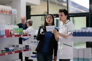 Pharmacist and medical store customer comparing vitamin boxes and reading usage instruction on packages. Pharmaceutical specialist helping young asian woman choosing between two multivitamin complex photo