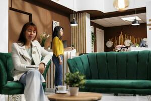 Asian tourist relaxing in hotel lobby. Happy customer sitting in hotel lounge. Female client savoring her coffee inside luxury hotel. Smiling woman enjoying her vacation in elegant hotel. photo