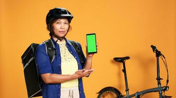 Woman holding chroma key green screen smartphone, showing location of upcoming delivery order. Courier preparing to ride bike to client address, isolated over orange studio background photo