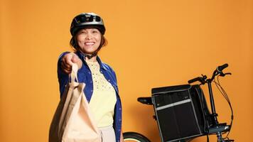 Courier waiting in front of apartment door for customer to pick up takeaway order. Food delivery cyclist offering takeout lunch bag to client, isolated over orange studio background photo