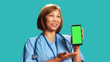 Professional hospital employee showing instructions video on phone green screen. Asian nurse wearing medical scrubs holding chroma key mock up phone, isolated over studio background, close up photo