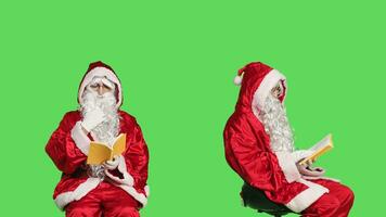 Father christmas with a reading hobby, enjoying lecture with poetry book or novel story while he sits on chair against greenscreen studio. Santa claus embodiment read literature or fiction. photo