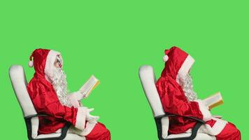 Saint nick reads poetry book on camera, enjoying literature or fiction lecture for education. Man dressed like santa sitting on chair with novel story in hand, greenscreen background. photo