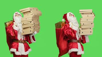 Santa delivering pizza boxes on greenscreen backdrop, carrying big pile of fast food and sack with toys for children. Saint nick in festive red costume acting like a deliveryman in studio. photo