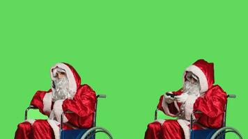 Man santa claus using smartphone, playing videogames and talking on phone call in studio. Person portraying father christmas in wheelchair with disability, having fun gaming and chatting. photo