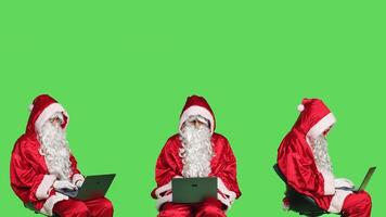 Modern santa claus works on laptop, greenscreen backdrop in studio. Father christmas sitting on chair and using wireless pc for online website browsing, searching for children addresses. photo