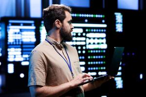 Proficient technician using laptop to check server hub devices. High tech rigs in data storage and cloud computing business service providing processing for several workloads photo
