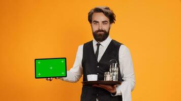 Smiling butler presents greenscreen on tablet, posing with confidence against yellow background. Young adult worker showing isolated template on smartphone display in front of camera. photo