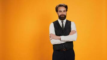 Refined waitstaff member poses happily in front of yellow background before bringing food dishes. Butler serving in formal environment while wearing suit in posh culinary industry. photo