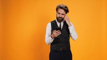 Before serving guests at table, classy butler checks phone looking at online web page against yellow background. Staff member with smartphone serving in an eatery, catering sector. photo