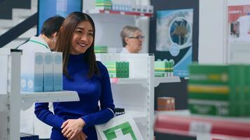 Smiling customer amidst drugstore shelves, looking at medicine pill packages, trying to find suitable remedy for troubling sickness. Happy cilent holding shopping bag, browsing through medicaments photo