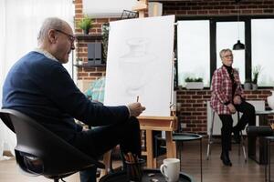 Senior man sitting at easel practicing drawing, visiting group art classes, learning new things in retirement. Mature male student sketching on canvas with pencil, attending creative arts activities photo