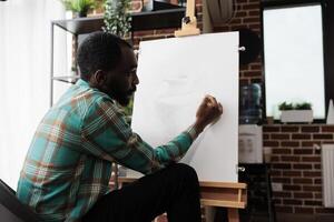 Young African American guy expressing himself through art, attending drawing classes, male student improving sketching skills. Talented artist sitting at easel creating masterpiece on canvas photo