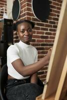 Arte clases y mental salud. africano americano estudiante niña sentado a caballete participación lápiz y sonriente a cámara mientras aprendizaje a dibujar, contento joven negro mujer expresando sí misma mediante dibujo foto
