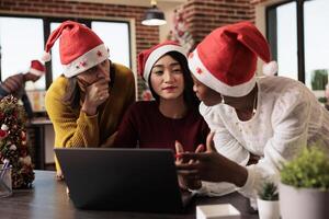 Women workers team gathered around laptop, discussing project tasks in office during christmas holiday. Diverse colleagues in santa hats collaborating on annual report on xmas eve photo