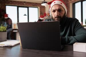 Focused employee in santa hat solving task problem while working on laptop in decorated festive office. Thoughtful company employee managing workload during xmas winter holiday photo