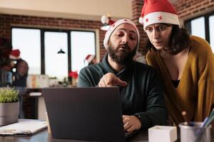 Company employee showing task problem on laptop to project manager while coworking in office at christmas eve. Coworkers in santa hats brainstorming and working at festive workplace photo