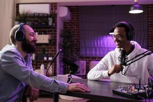 Online show host recording podcast with guest, using audio recording equipment in home studio living room. Happy men laughing together during broadcasting session for internet streaming service photo