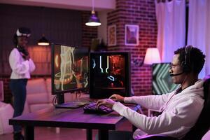 Man immersed in gaming session at computer desk while girlfriend in background uses virtual reality goggles. African american couple playing games, having fun on gaming pc and vr system photo