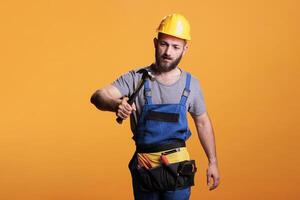 Construction worker using hammer on renovation project, standing over yellow background. Professional male renovator doing reconstruction work with sledgehammer and tools, studio shot. photo