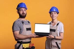Team of builders showing white empty screen on laptop, wearing overalls and hardhats in studio. Construction workers advertising isolated copyspace display on portable computer, blank mockup. photo