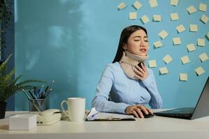 Asian woman with cervical collar on neck using laptop computer feeling discomfort and pain. Female corporate employee with injury working seated at startup company workstation. photo