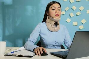 Corporate employee wearing medical collar for injury while working on reports, preparing paperwork for next meeting. Injured female entrepreneur concentrating on finishing project at work station. photo