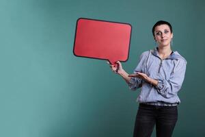 Cheerful model pointing at empty red speech bubble in studio, advertising copy space accessory for text message. Smiling attractive woman looking at camera while showing blank template board photo