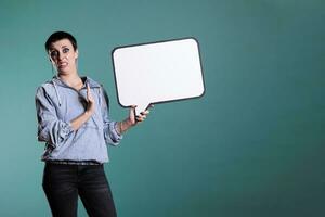 Cheerful brunette woman pointing at empty white speech bubble during studio shot, advertising copy space accessory for text message. Smiling model looking at camera while showing blank template board photo