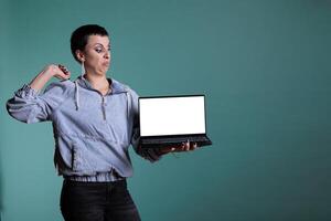 Angry unhappy woman with disapproval expression presenting laptop computer with blank screen for copyspace in studio. Sad negative female doing dislike gesture standing over isolated background. photo