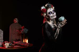 Spooky santa muerte model posing with roses over black background, looking like goddess of death. Beautiful woman with skull make up and body art, festival holiday celebration in studio. photo