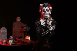 Creepy lady of death with skull make up and colorful wreath of flowers, acting like holy santa muerte on day of the dead celebration. Beautiful goddess wearing festival tradition costume. photo