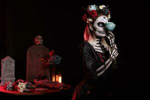 Glamour woman dressed as catrina skull in studio, wearing traditional santa muerte holy costume with black and white make up or body art. Holding roses and having flowers crown. photo