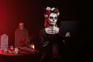 Model with halloween costume holding laptop, posing on tranditional mexican holiday celebration. Wearing skull make up and day of the dead costume with body art, goddess of death. photo