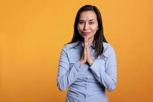 positivo asiático mujer Orando a Dios en frente de cámara, participación manos en oración gesto y preguntando para bueno suerte. religioso modelo expresando esperanza y creencia, en pie en estudio terminado amarillo antecedentes foto