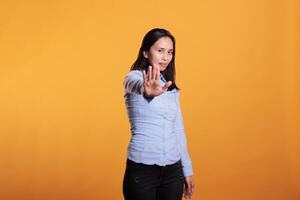 Asian positive woman doing stop symbol with palm on camera, expressing disapproval and denial. Cheerful model showing rejection and refusal gesture standing in studio over yellow background photo