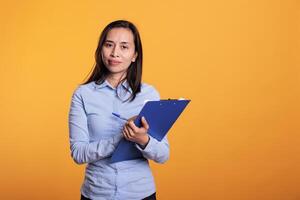 asiático mujer tomando notas en azul portapapeles con bolígrafo en estudio. pensativo filipino persona escritura reporte en cuaderno archivos, trabajando a negocio proyecto respondiendo cuestionario pregunta foto